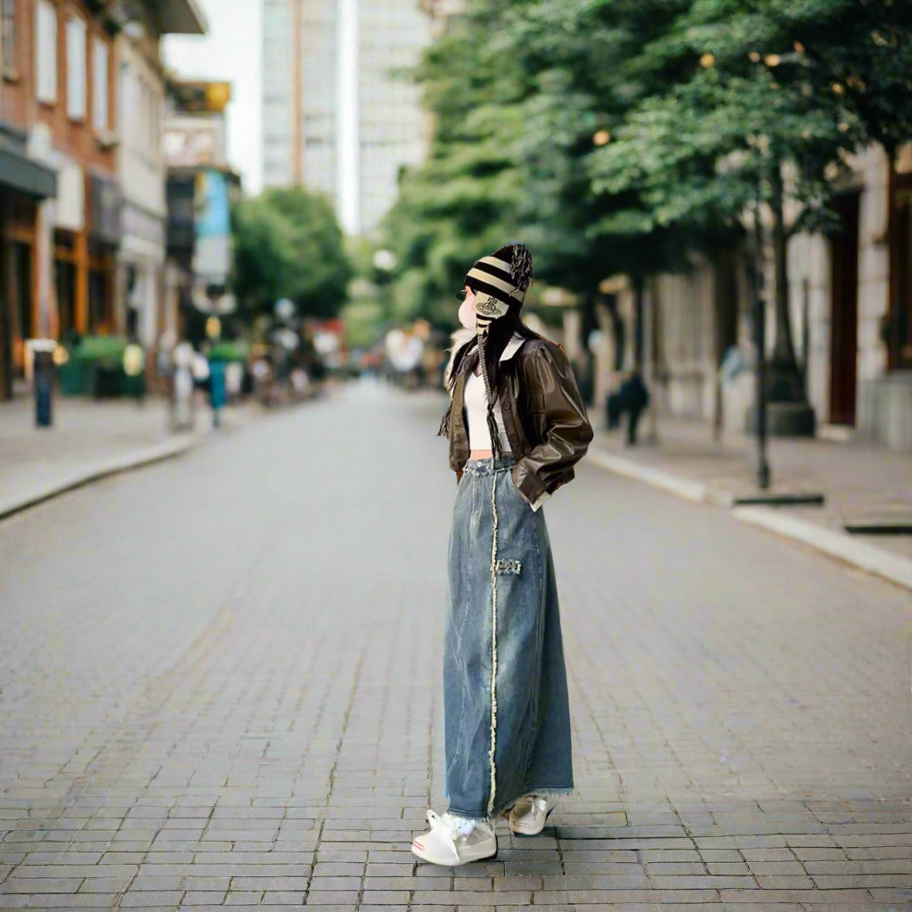 Person wearing a brown leather jacket, striped beanie, and blue jeans standing on a sidewalk.